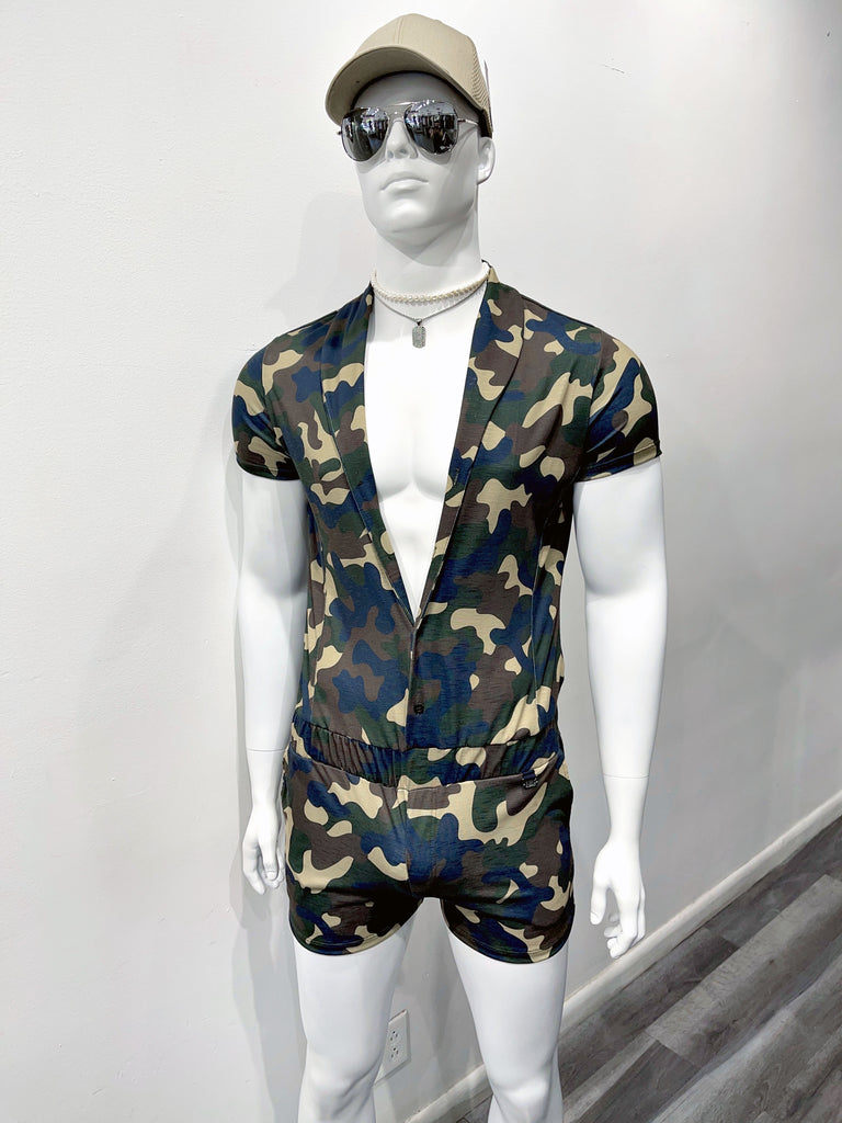 A mannequin in dark mirrored sunglasses, a tan baseball cap, a thin silver choker chain with fake diamonds, and a thin silver chain with a silver dog tag covered in fake diamonds, and a green camouflage shorts romper that is unbuttoned down to his waist.