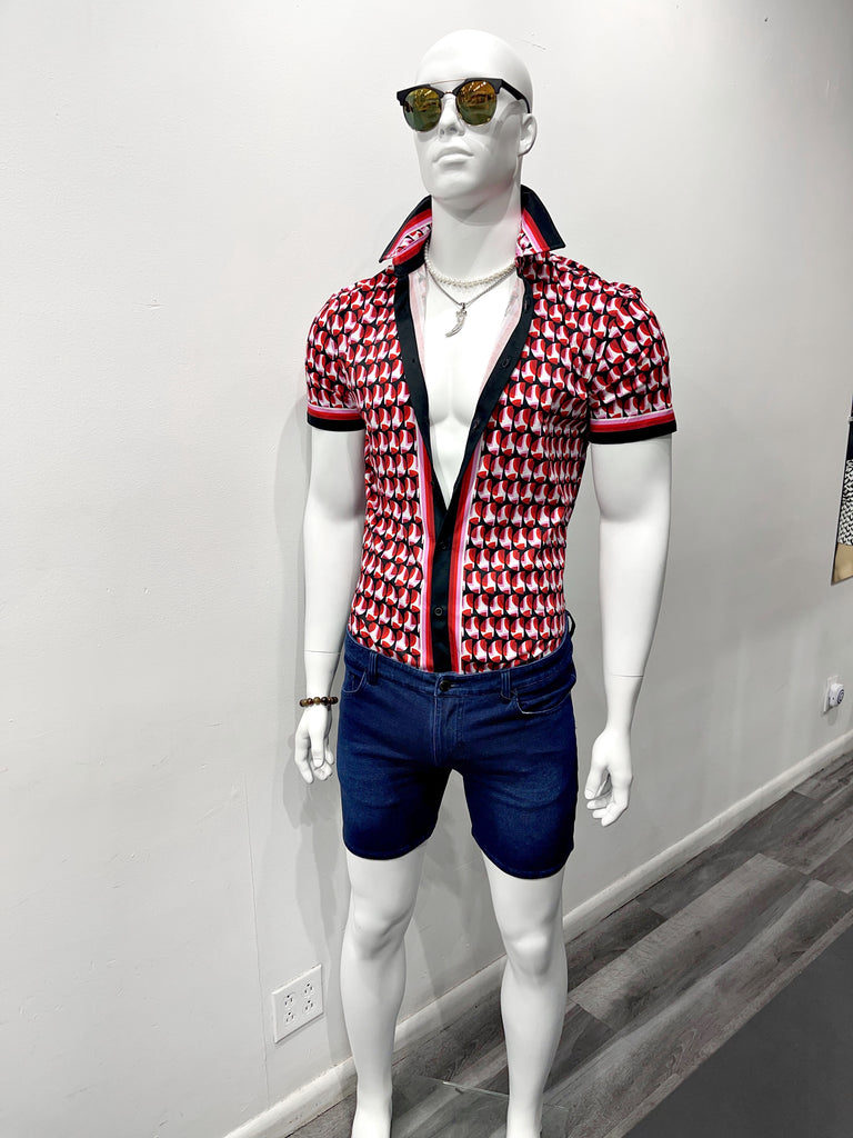 A white mannequin wearing colorful mirrored sunglasses, a gold choker chain, a silver chain necklace with a shark tooth pendant, a short-sleeved button-down shirt with a bold red, pink and navy-blue mod pattern, and blue stretch denim shorts.