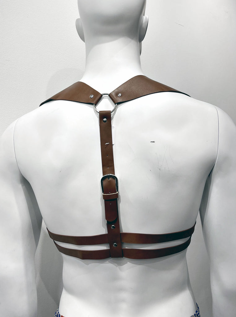 Brown harness as seen from the back, comprised of thick shoulder straps that both connect to a single silver ring in the mid back, which connects to a single thin strap that goes down the back and wraps around the two torso straps that encircle the mid-torso horizontally.