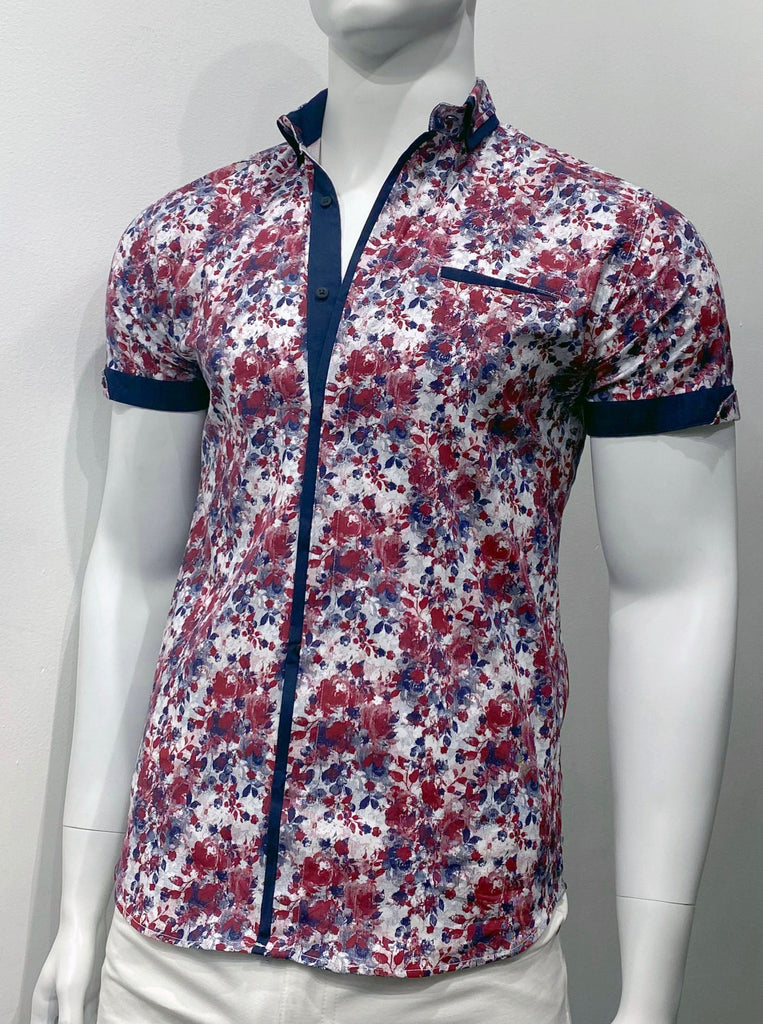 Short-sleeved button down shirt with a navy blue and burgundy floral pattern on off-white fabric, as seen from the front. The buttons are dark, shiny gunmetal. The sleeve cuffs, the inside of the collar, and the placket are navy blue fabric. There are button details on the sleeve cuffs and buttons on each collar. There is a left breast pocket with navy blue fabric detail poking out of the top.
