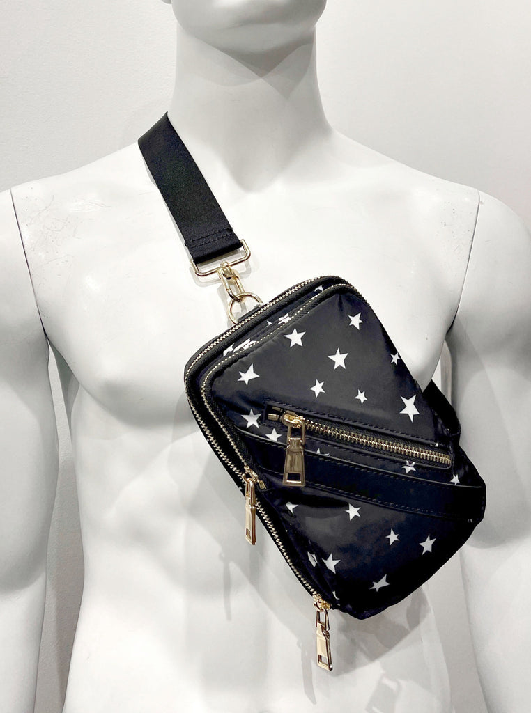 Small, black sling backpack that has 2 zipper compartments and 1 front zip pocket with gold zippers. The bag has a black and white striped shoulder strap, vegan leather detailing on the bag, and a pattern of white stars all over it.