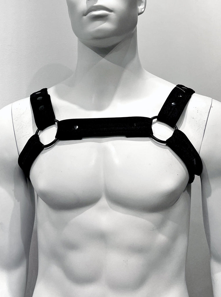 Black chest harness as seen from the front. It has a thick, black strap that encircles the chest comprised of three shorter, equally thick straps—one that goes across the chest, and two that go under each arm—all connected together by black metal rings atop the left and right breasts. Two more straps go over each shoulder, also connected to the large, metal rings. All of the straps line of small, black metal circles running lengthwise down the center of the strap.