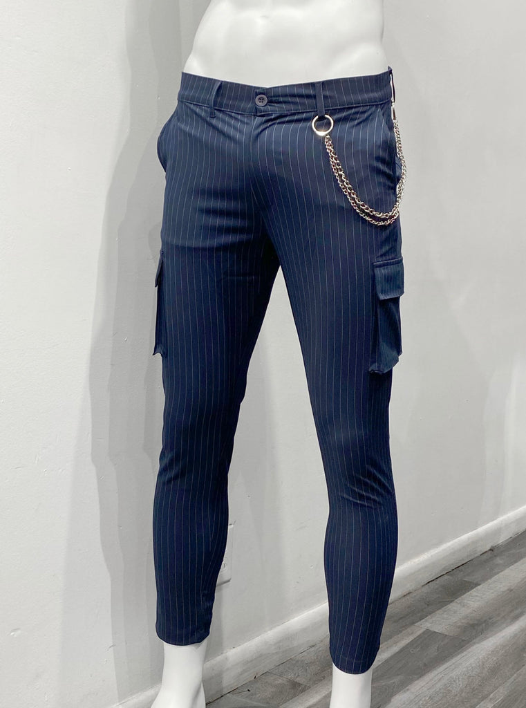 Navy blue slim fit, flat-front pants with white vertical pin stripes as seen from the front. There is a single utility cargo pocket on the outside of each leg. There is a silver fashion chain draped over the left hip, hanging from a left front belt loop and a left back belt loop.