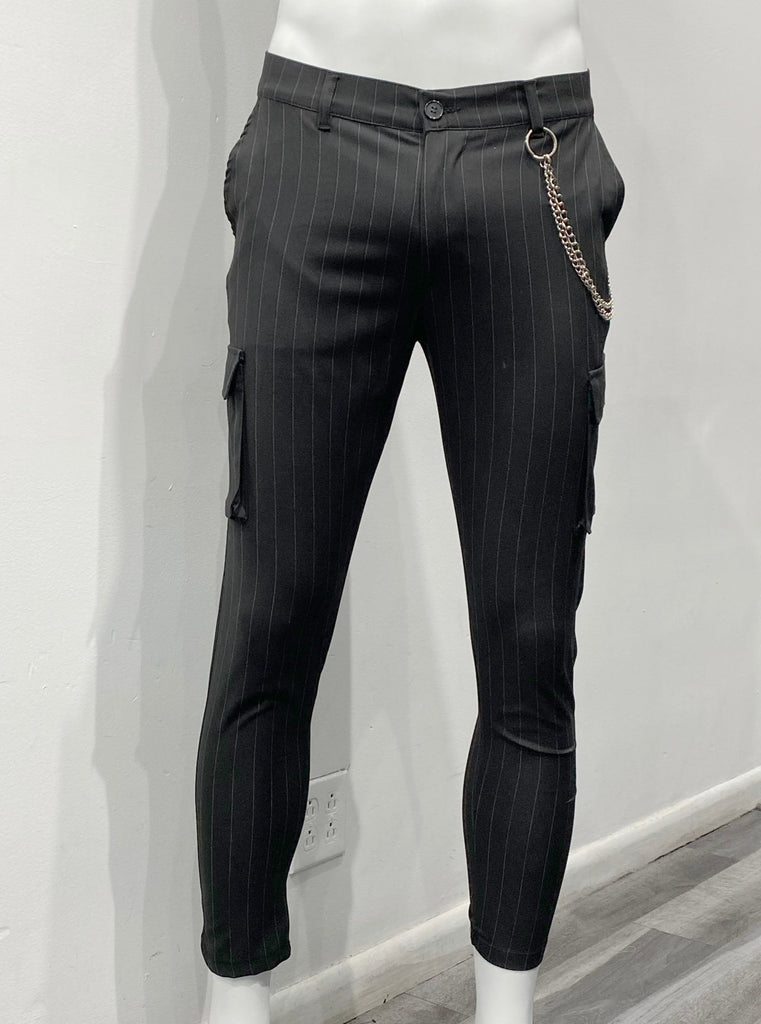 Black slim fit, flat-front pants with white vertical pin stripes as seen from the front. There is a single utility cargo pocket on the outside of each leg. There is a silver fashion chain draped over the left hip, hanging from a left front belt loop and a left back belt loop.