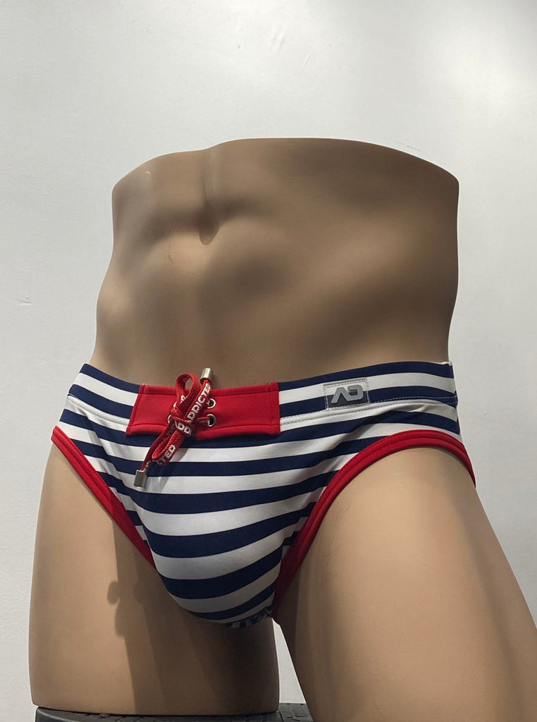 White swim brief as seen from the front, with horizontal blue stripes. There is a red rectangular patch front and center on the waistband, in the drawstring closure area, with a grey brand emblem on the waistband above the left hip. Also there is red piping around both leg openings.