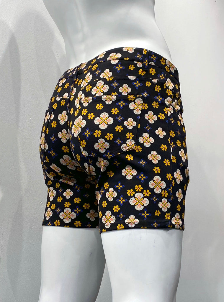 Zip-front, 5-pocket black stretch knit shorts as seen from the back, covered with a graphic floral pattern comprised of flowers with white petals, orange centers and pink accents, smaller orange flowers, and ever smaller orange and violet flowers.
