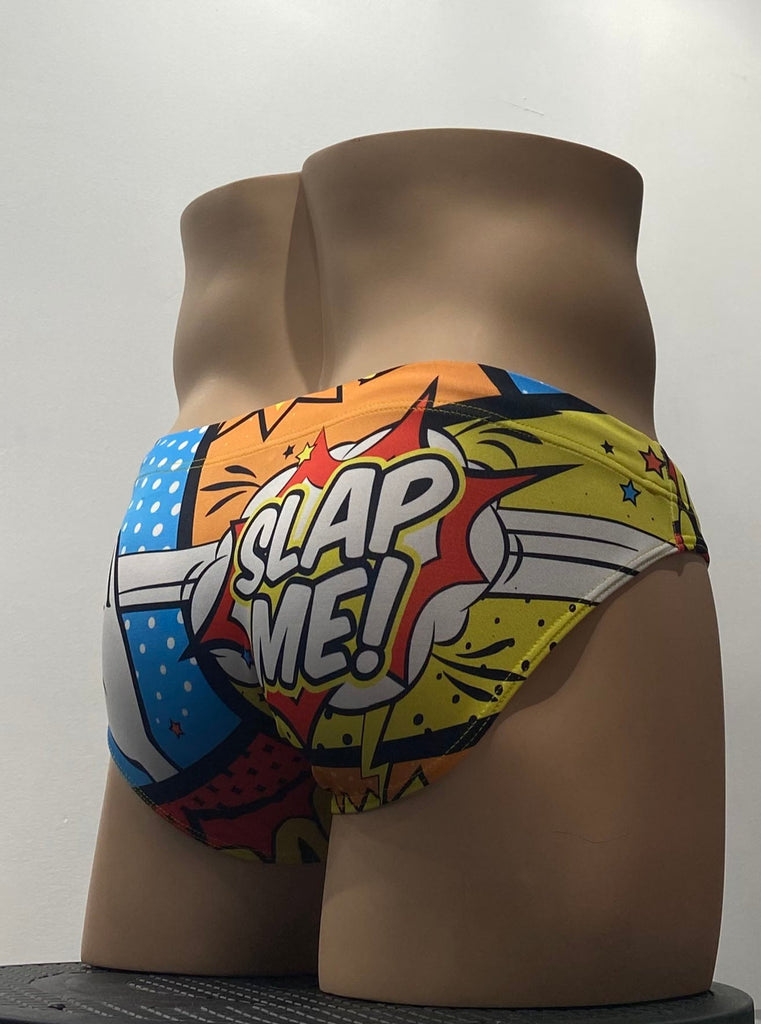 Multi-color swim brief with a yellow, blue, orange, red, and black pop art design as seen from the back. The words “Slap me” are on the butt with the rendering of a white cartoon hand slapping the back of the suit.