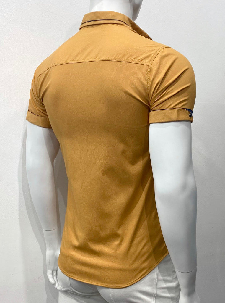 Mustard yellow short-sleeved button down shirt as seen from the back. The back side of the navy blue sleeve cuffs are covered with an additional cuff of the mustard yellow fabric that encircles just the back side of the arm holes and covers up most of the navy blue arm cuffs, so just a sliver of navy blue is visible over the mustard yellow cuffs. There is a subtle paisley pattern woven into the mustard yellow fabric. The fabric on the inside back of the collar is navy blue and it peeks out under the collar.