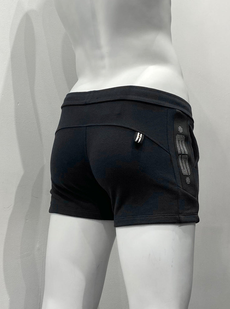  Black athletic shorts as seen from the back, with a white and black striped tag detail with a silver gromet in it on the back, lefthand side of the shorts. On the outside of the right hip, there is a thick, black vertical strip with a brand emblem comprised of many small, shiny metal circles.