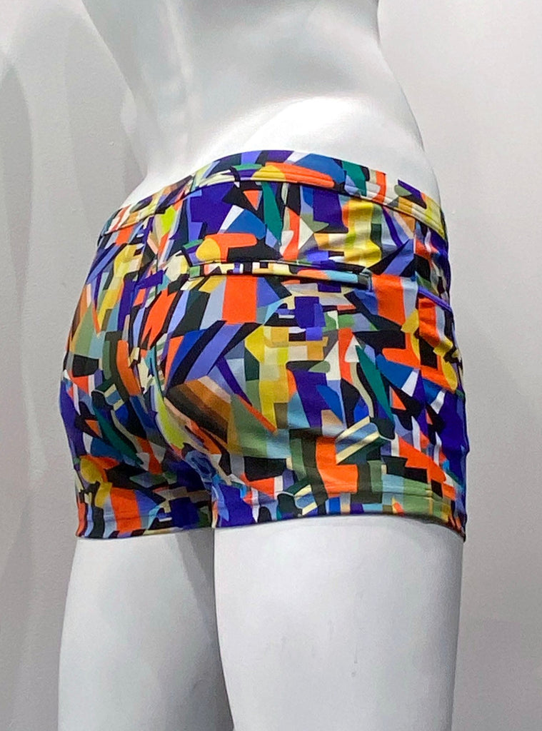 Swim shorts with a 3-inch inseam, front pockets as seen from the back. The shorts are covered with a vibrant multicolor angular prism pattern comprised of many different shades of purple, orange, blue, green, yellow, brown, grey, with black. There is a hidden zip pocket on the back, right seat.