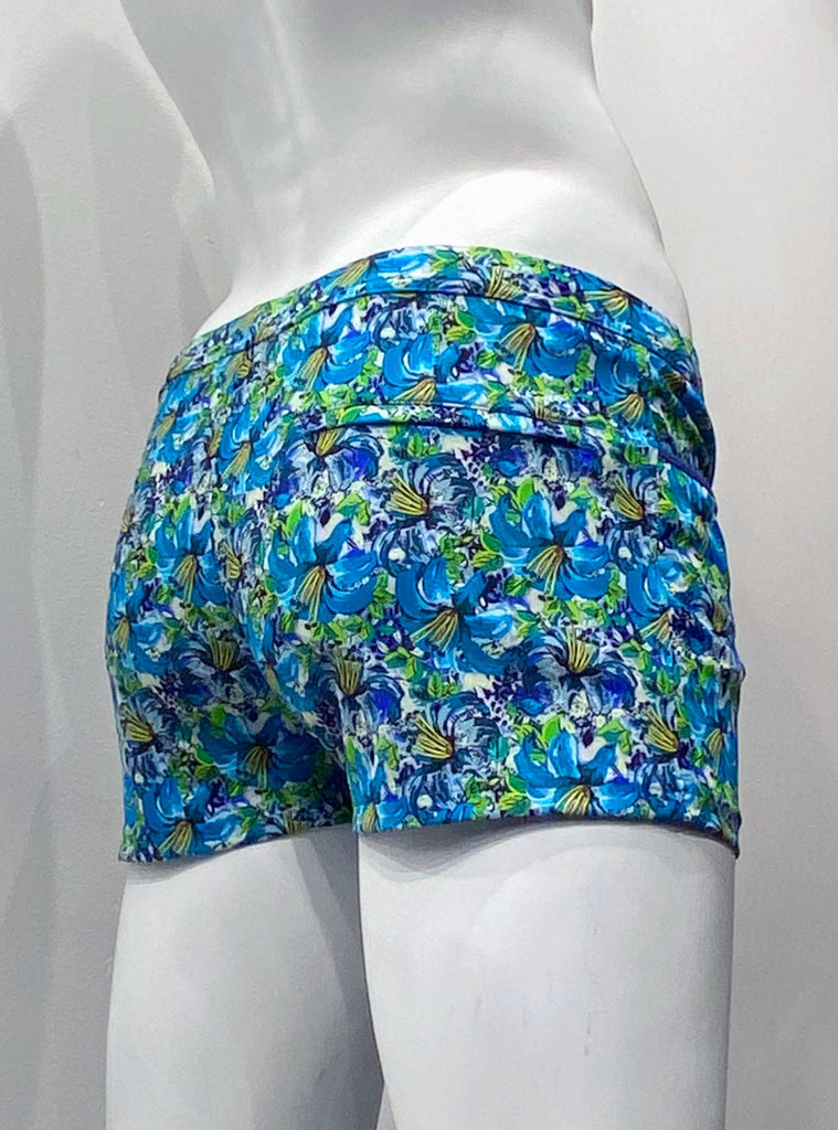 Swim shorts with a 3-inch inseam, front pockets as seen from the back. The shorts are covered with a teal, blue, yellow, and light green floral pattern. There is a hidden zip pocket on the back, right seat.