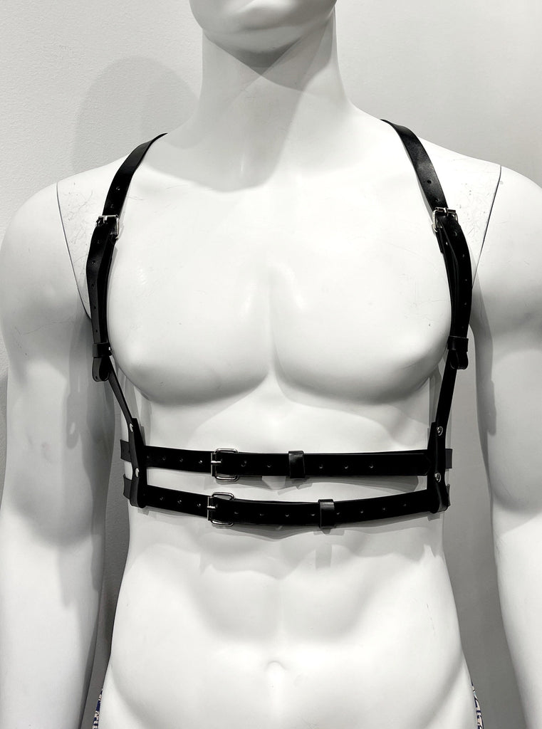 Black vegan leather harness as seen from the front. A single, thin shoulder strap goes over each shoulder, extending down the front of the torso on each side. These shoulder straps are each made of two shorter straps that are attached together by a sliver belt buckle. Each shoulder strap reaches down on either side of the chest and wraps around two equally thin torso straps that encircle the mid-torso horizontally and buckle in the front.